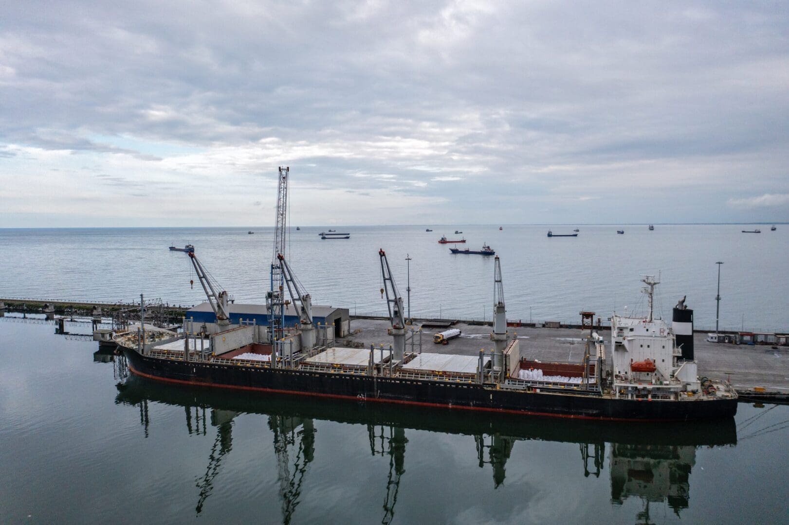 Large cargo ship docked at a harbor with cranes and cloudy sky.